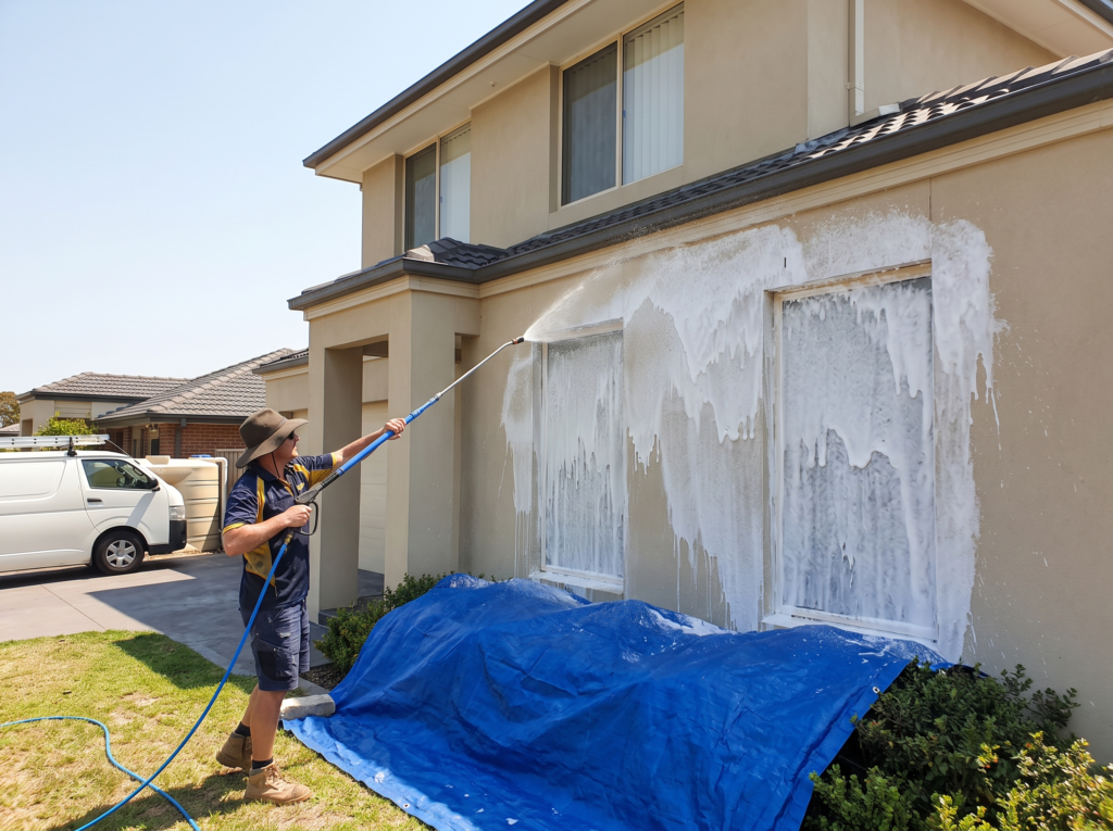 Technician soft washing a rendered house wall with low pressure foam and protected garden below