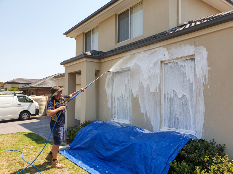 Technician soft washing a rendered house wall with low pressure foam and protected garden below